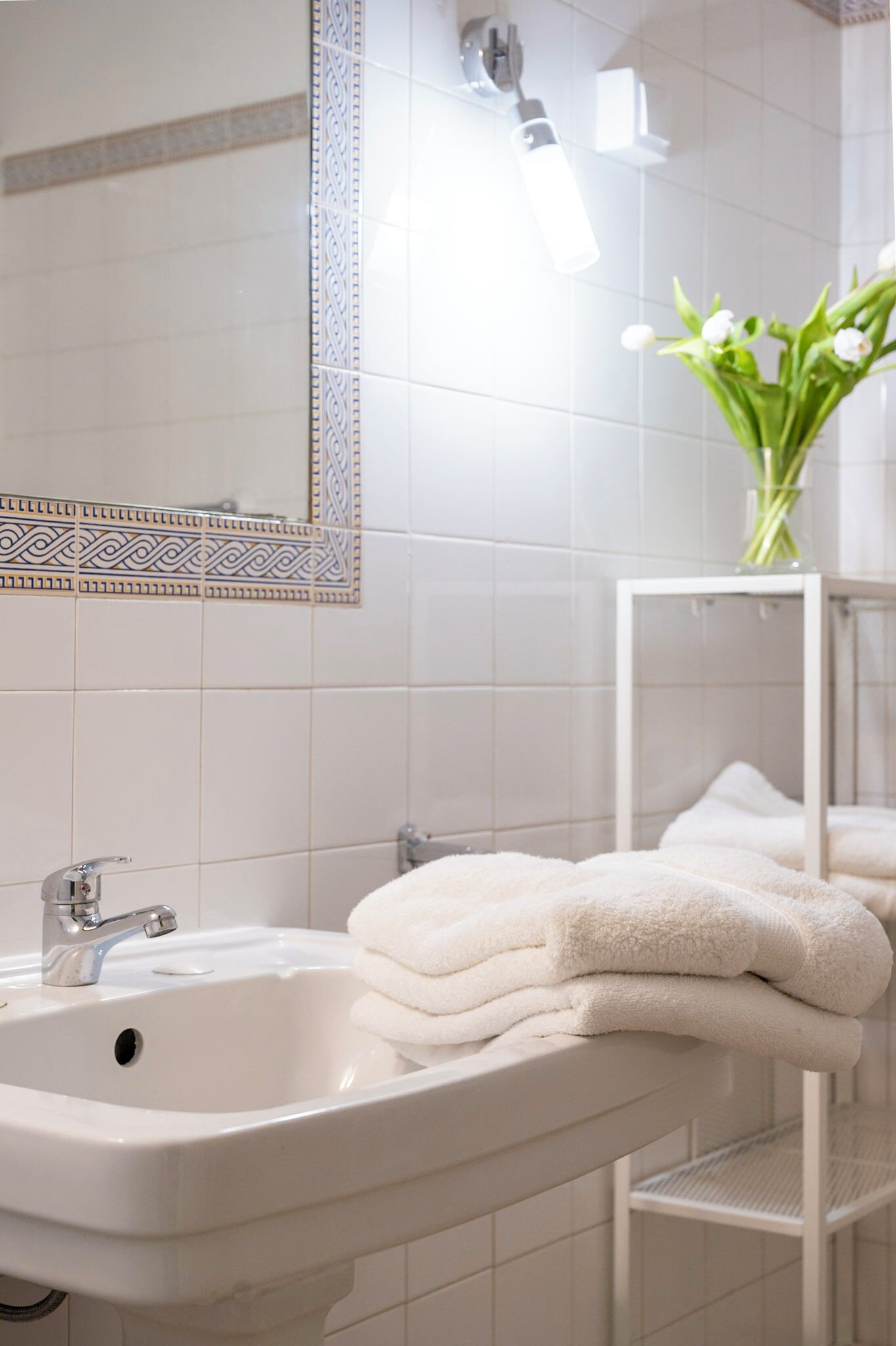 Un baño con un lavamanos blanco, un espejo con azulejos decorativos y un perchero con toallas dobladas.
