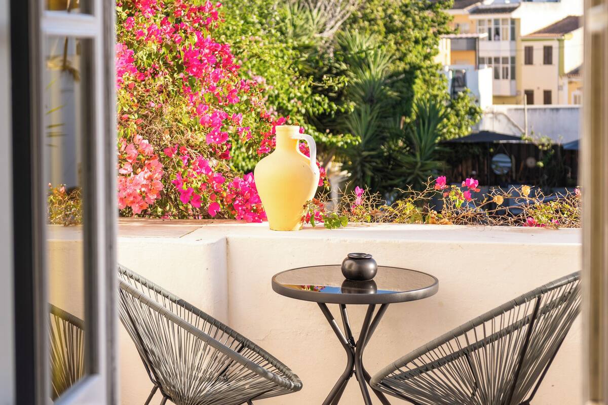 Un balcon ouvert avec deux chaises en fil de fer, une petite table ronde et un vase jaune.