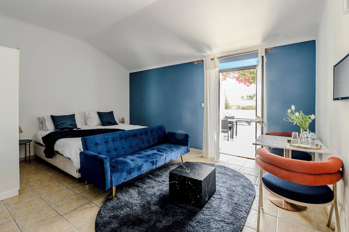 Une chambre moderne avec un canapé bleu, un pouf noir et un coin repas avec des chaises orange.