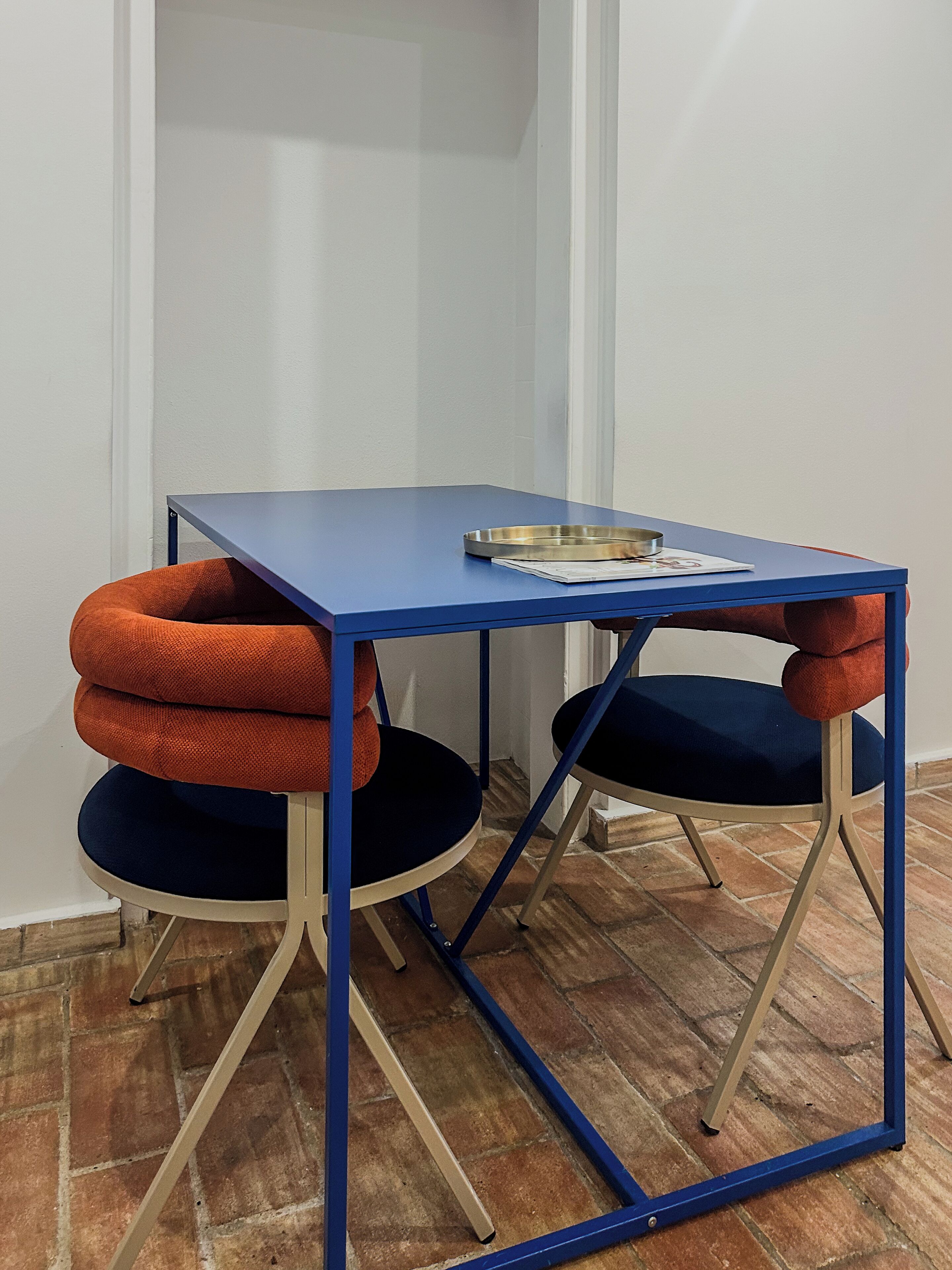 A small, blue table with a metal tray and papers, flanked by two chairs with orange and blue cushions.