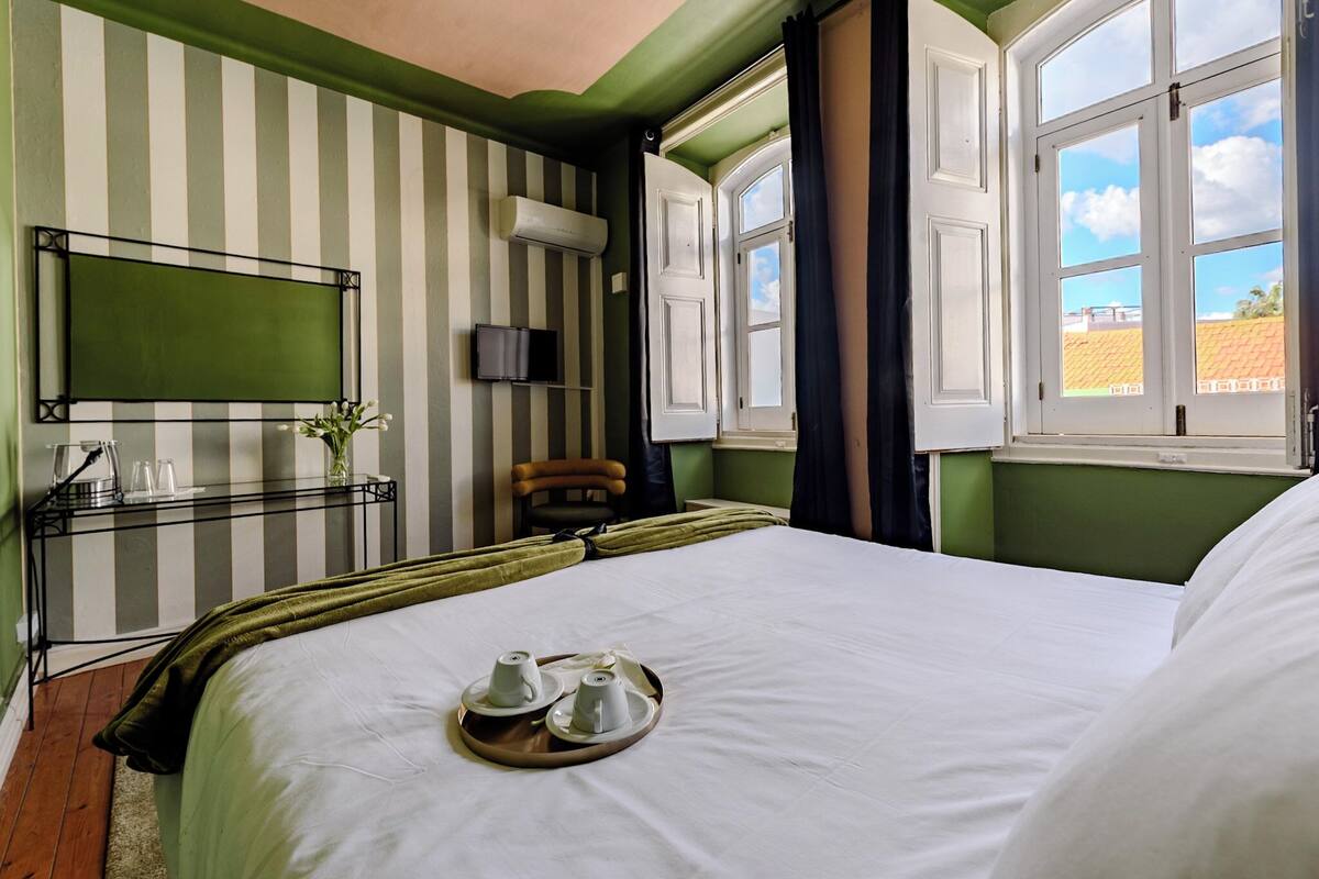 Une chambre d’hôtel avec un lit, une table de chevet sur laquelle se trouve une assiette avec des tasses et des sous-tasses, un téléviseur posé sur un meuble, une fenêtre avec des rideaux et un mur à rayures.