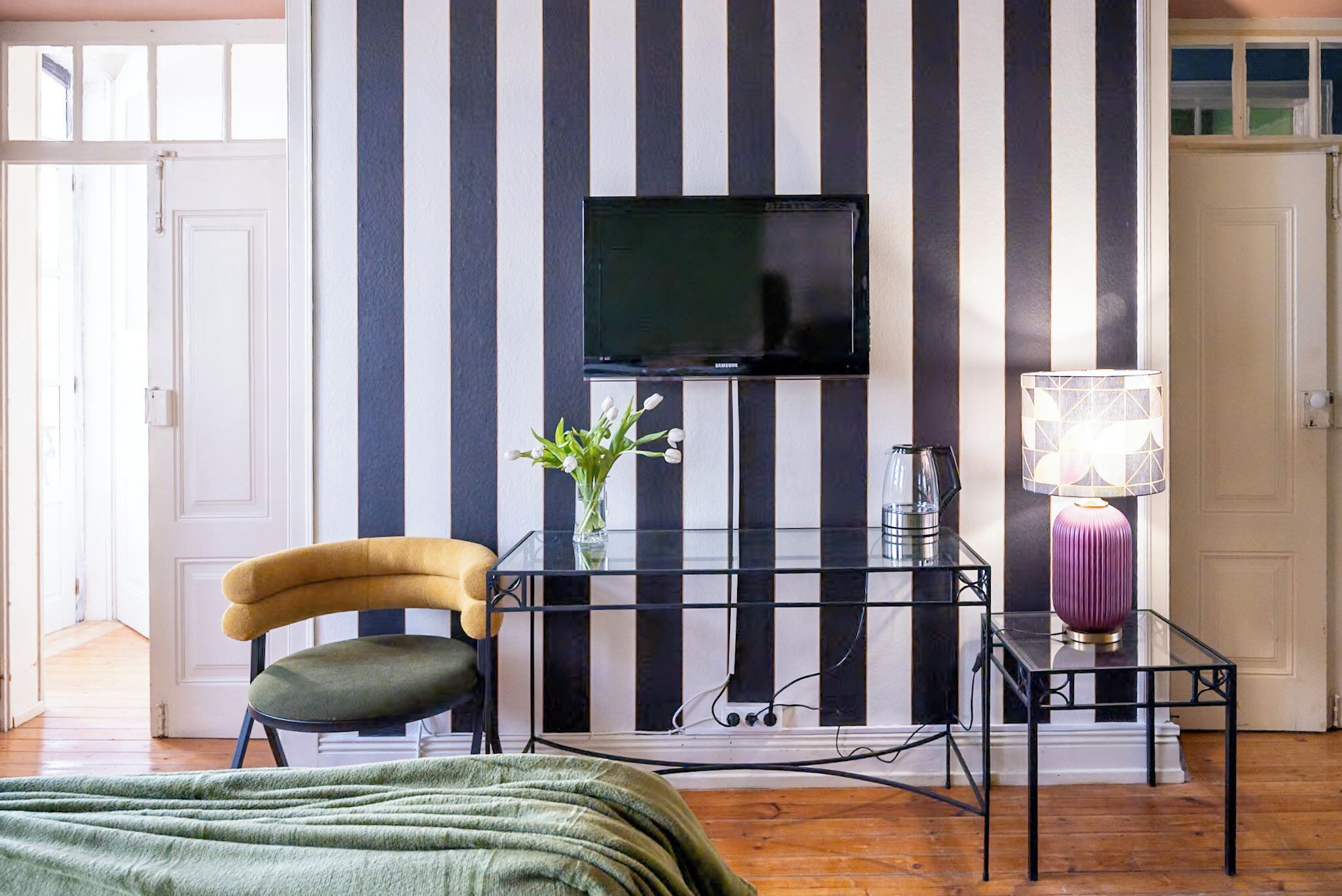Une pièce avec un mur à rayures, un téléviseur fixé au-dessus d’une console noire, une chaise et une lampe.