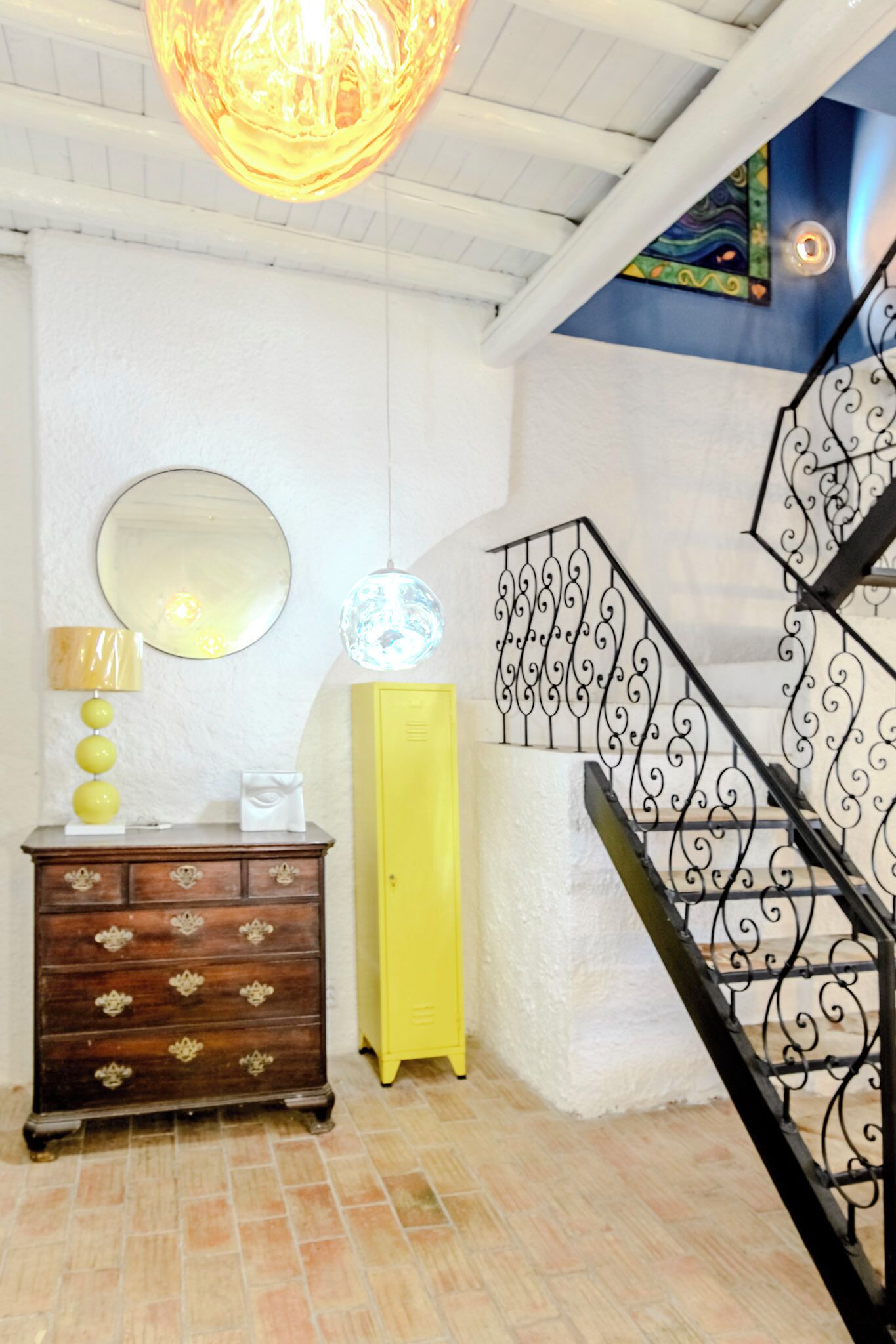 A staircase with ornate black railings, a yellow cabinet, a wooden dresser with decorative handles, a round mirror, and a hanging light fixture.