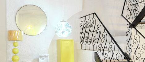 A staircase with ornate black railings, a yellow cabinet, a wooden dresser with decorative handles, a round mirror, and a hanging light fixture.