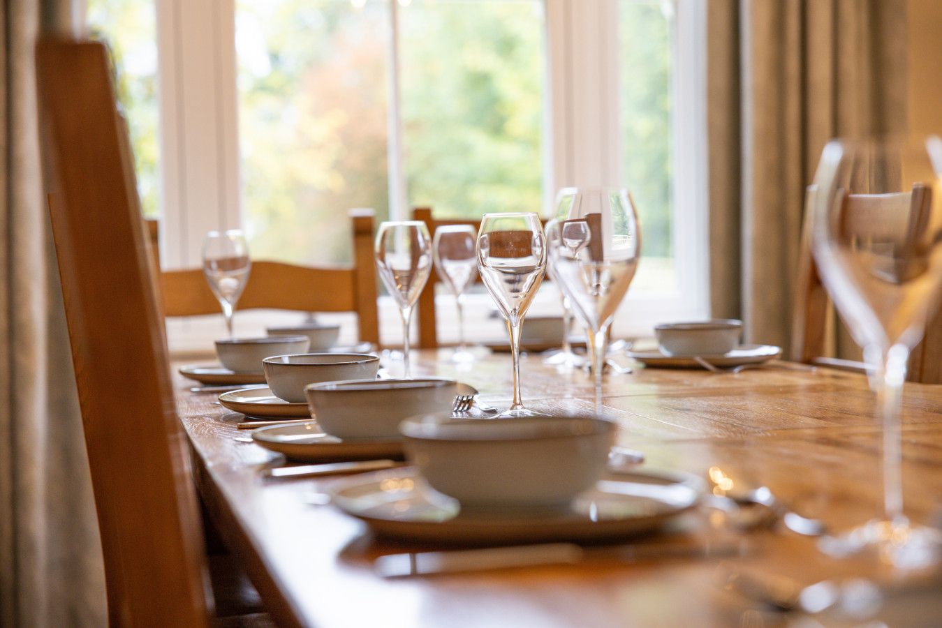 A dining table set with multiple wine glasses and bowls, with a view of greenery through the window.