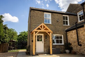 A two-story brick house with a wooden front porch, a white door, and a small garden area with potted plants. - The Maid's Cottage  · Rural hideaway in a cosy farm cottage with hot tub (Sawtry)