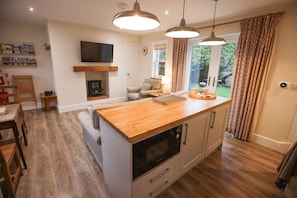 A modern kitchen with a central island, wooden flooring, pendant lighting, a built-in oven, and a television on the wall. - The Maid's Cottage  · Rural hideaway in a cosy farm cottage with hot tub (Sawtry)