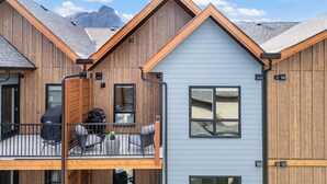 Ein modernes, mehrstöckiges Gebäude mit Holzfassade, einem Balkon mit Gartenmöbeln und Bergblick im Hintergrund.