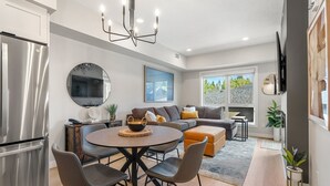 Eine moderne Küche mit einem Edelstahlkühlschrank, eine Essecke mit rundem Tisch und Stühlen, ein großer Spiegel und ein Wohnbereich mit Sofa und Wand-Fernseher.