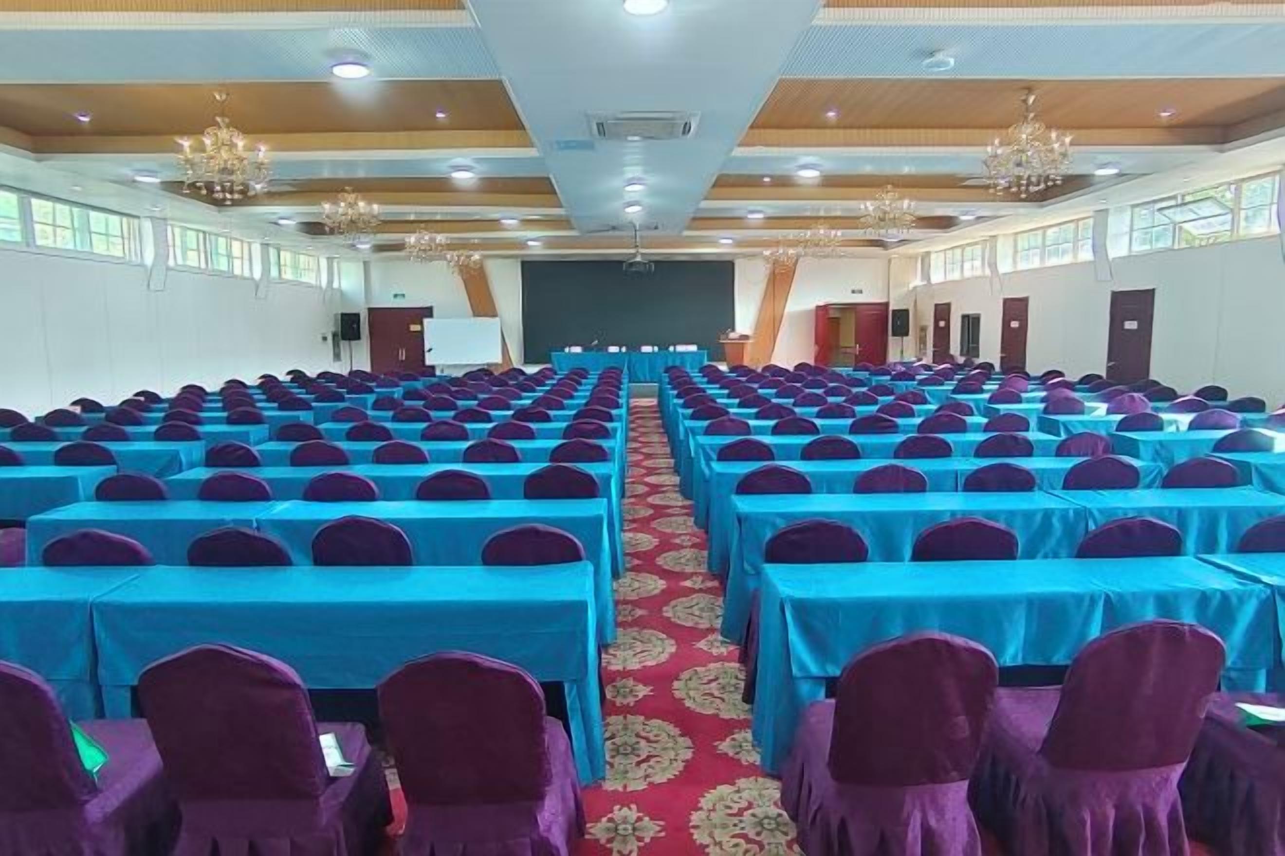 A conference room with rows of tables and chairs, a stage at the front, and a chandelier.