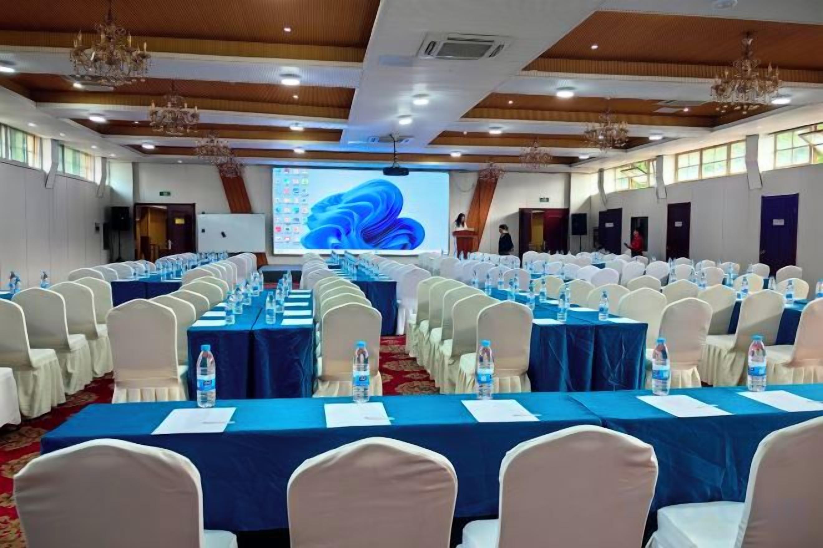 Un salón de conferencias con filas de mesas, sillas y una pantalla grande al frente.