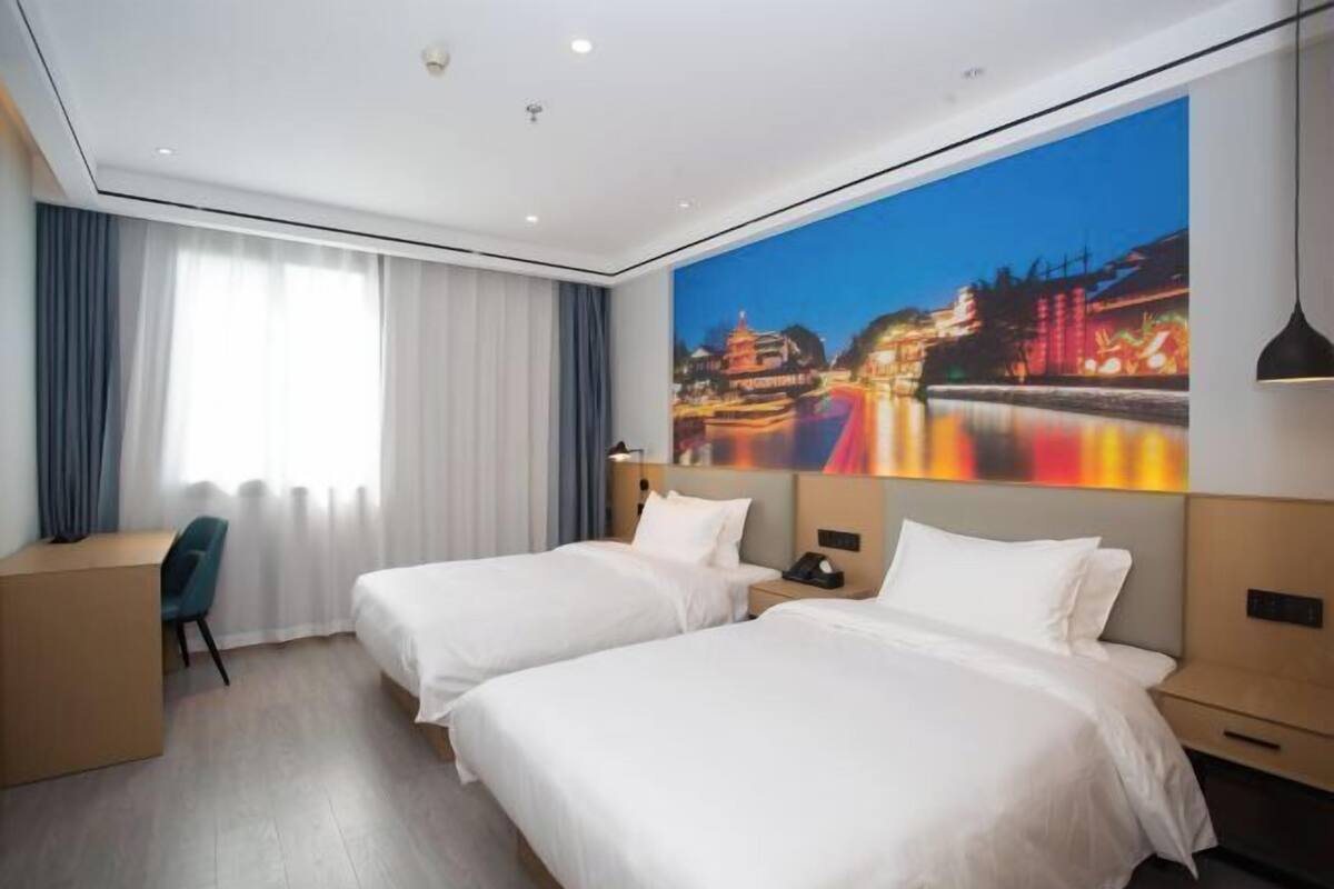 Habitación de hotel con dos camas, un escritorio, una silla, un ventanal con cortinas y un cuadro de un paisaje urbano en la pared.