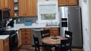 A compact kitchen with wooden cabinets, a stainless steel refrigerator, a microwave, and a dining area with a table and chairs. - Eagle’s Landing (Lock Haven)