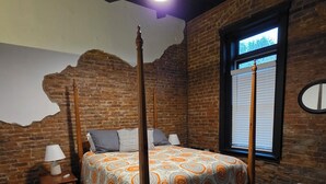 A four-poster bed with a patterned bedspread, a nightstand with a lamp, a window with blinds, and a brick wall. - The Delaney's Saloon (Vevay)