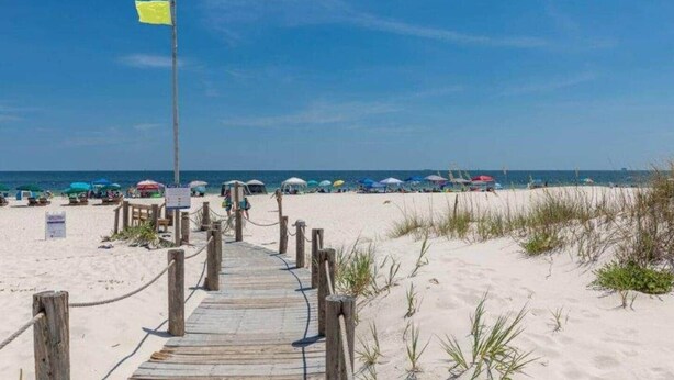 Ein hölzerner Steg führt zu einem Strand mit Sonnenschirmen und einer Flagge.