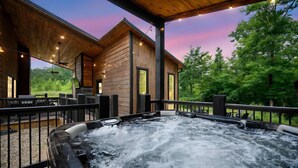 A hot tub on a wooden deck with a view of a cabin and trees. - Bearly Creekin' Stay for 14! (Broken Bow)
