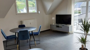 A modern living room with a dining area, a flat-screen TV, and a large window offering a view of the outdoors.