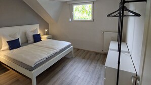 A bedroom with a bed, a bedside table, a hanging clothes rack, and a window with a view of greenery.