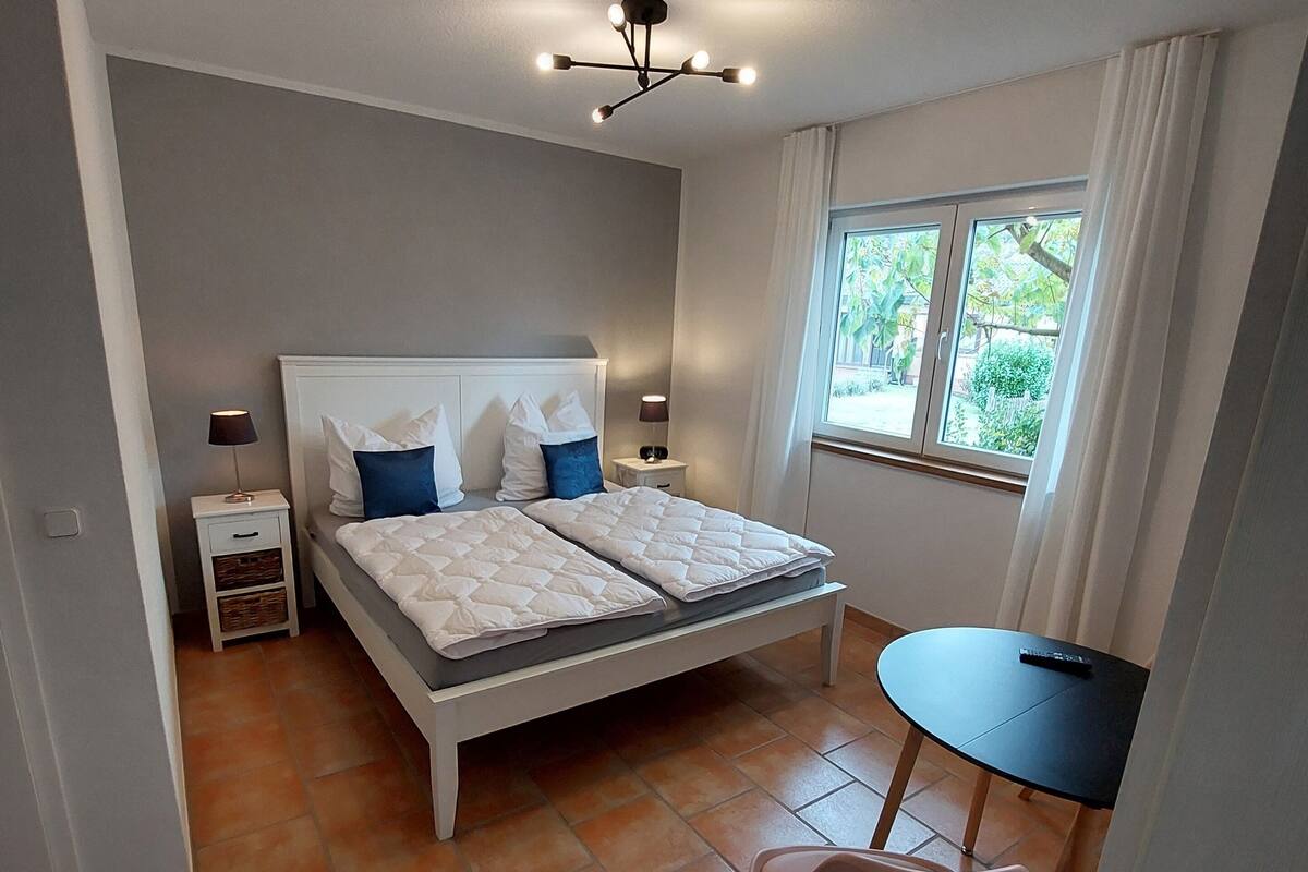Un dormitorio con cama, mesitas de noche, una silla, una pequeña mesa y una ventana.