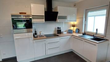 A modern kitchen with white cabinetry, a built-in oven, a black range hood, and a window with blinds.