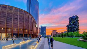 Una plaza urbana moderna con un edificio curvo, un rascacielos alto y una estructura vertical singular con vegetación.