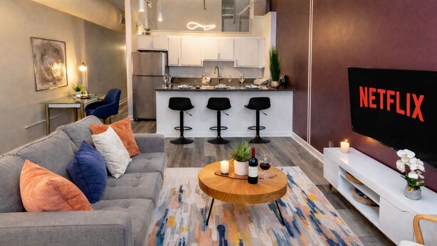 Un salon moderne avec un canapé gris, une table basse ornée de bougies et une cuisine en arrière-plan.