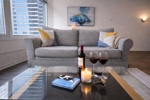 Una sala de estar moderna con un sofá gris, una mesa de centro de cristal, un libro, una vela y copas de vino.