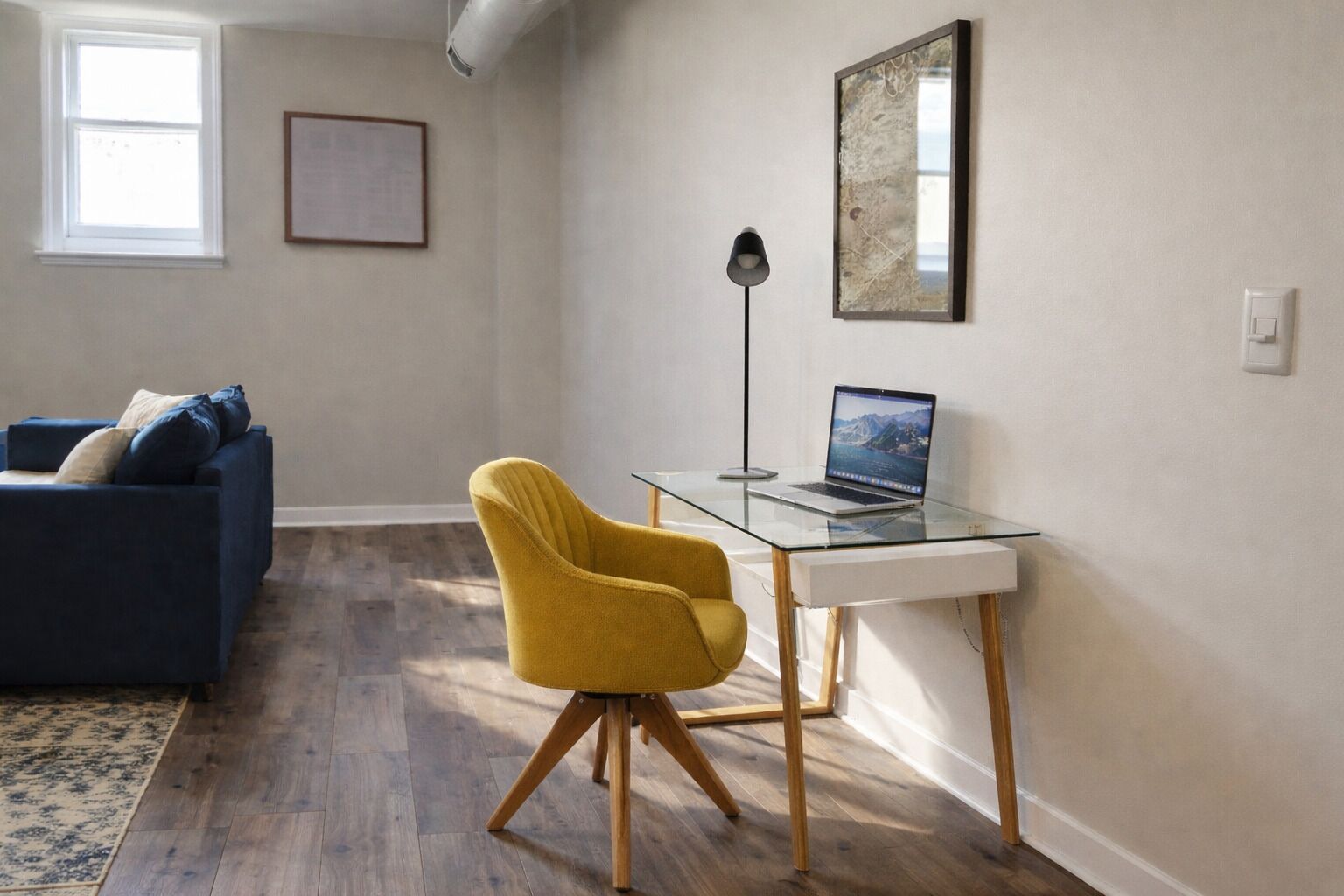A modern living room with a sofa, a yellow chair, a glass desk with a laptop, a lamp, and a framed picture on the wall.