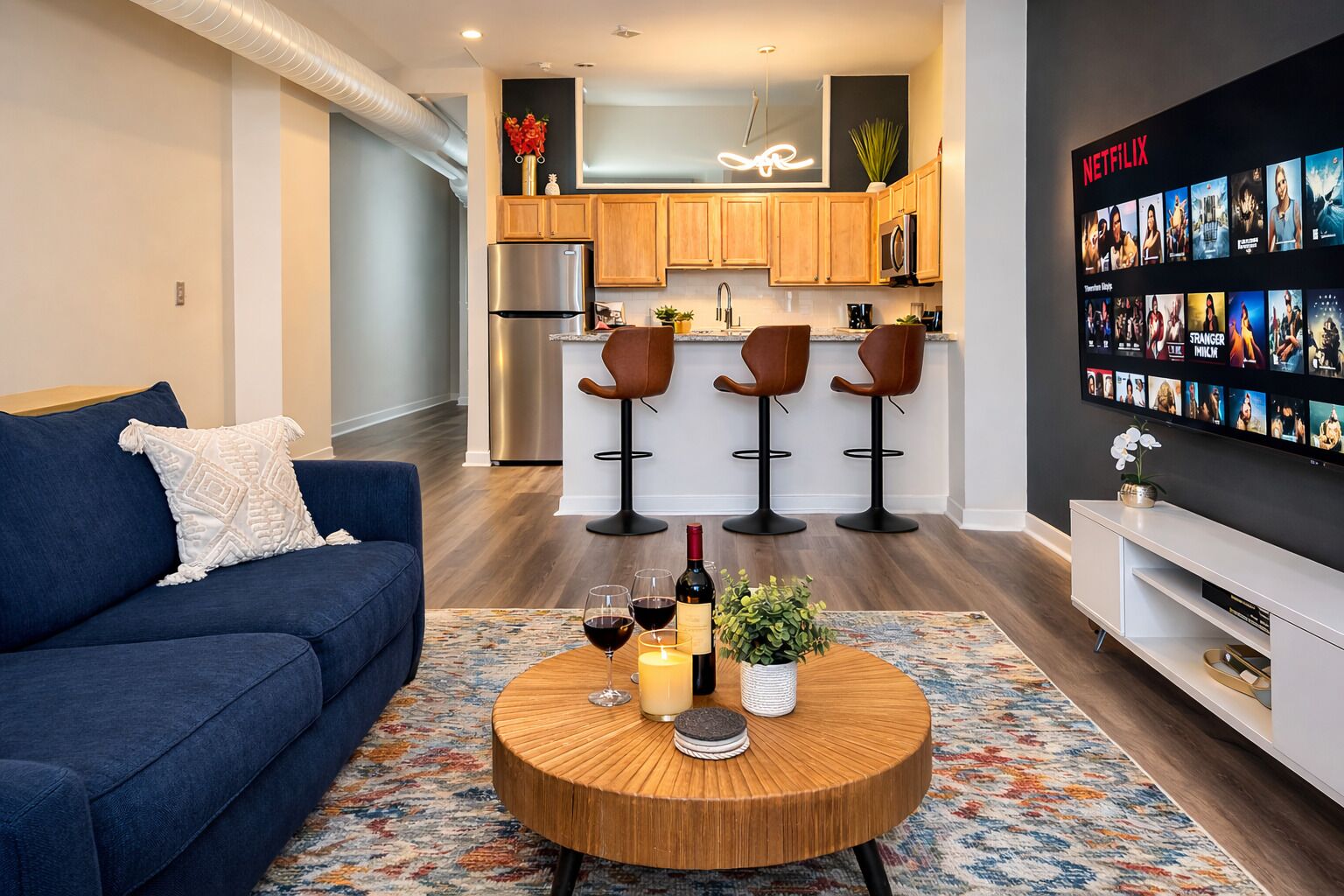 Un soggiorno moderno con un divano blu, un tavolino rotondo con bevande e una cucina con pensili in legno e una TV che mostra Netflix.