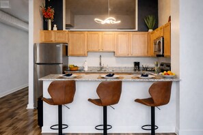 A modern kitchen with wooden cabinets, stainless steel appliances, a marble countertop, and three bar stools. - Ste 625+Skywalk Access to Convention Center+Free Parking (Indianapolis)