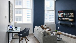 A modern living room with a sofa, a desk with a laptop, a TV mounted on the wall, and large windows.