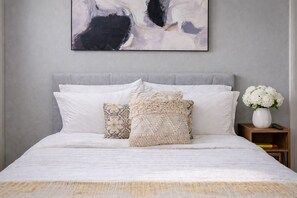 Un lit bien fait, avec une literie blanche et des coussins décoratifs, une tête de lit grise et un tableau abstrait accroché au mur.