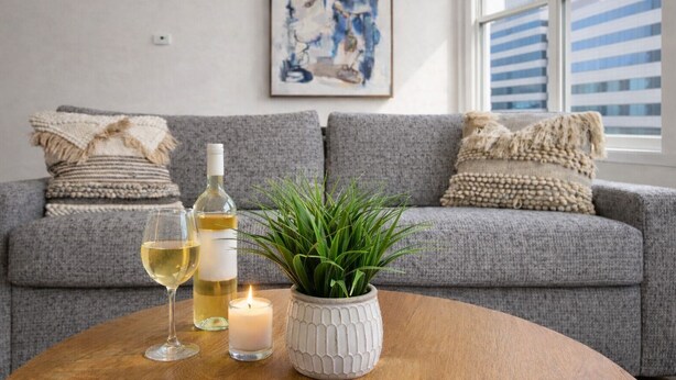 A grey sofa with decorative pillows, a bottle of white wine, two glasses of wine, a lit candle, and a green plant on a wooden table.