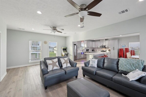 A modern living room with two grey sofas, a ceiling fan, and a kitchen in the background. - Shore Thing - Beach Front - Sleeps 22 (Port Bolivar)