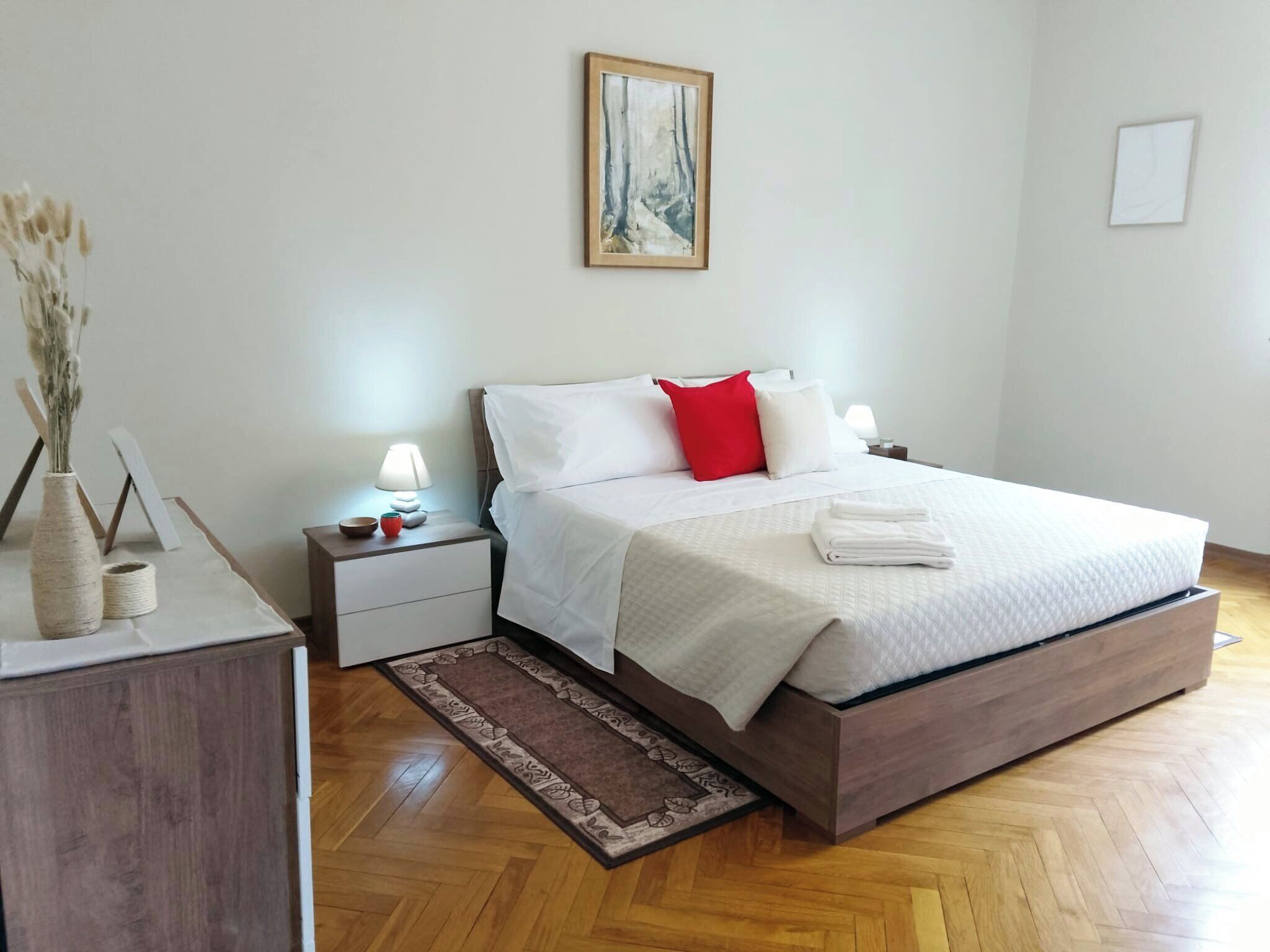 A neatly arranged bedroom with a bed, bedside tables, a nightstand, a vase with dried flowers, and a painting on the wall.
