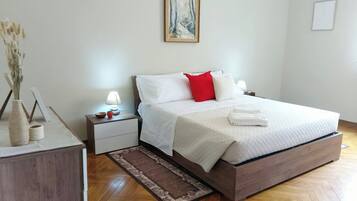 A neatly arranged bedroom with a bed, bedside tables, a nightstand, a vase with dried flowers, and a painting on the wall.
