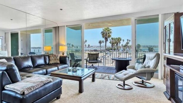 A spacious living room with a black leather sofa, a glass coffee table, and a view of palm trees and a body of water through large windows.