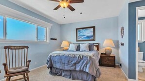 A bedroom with a bed, a chair, a ceiling fan, a window, and a painting on the wall.