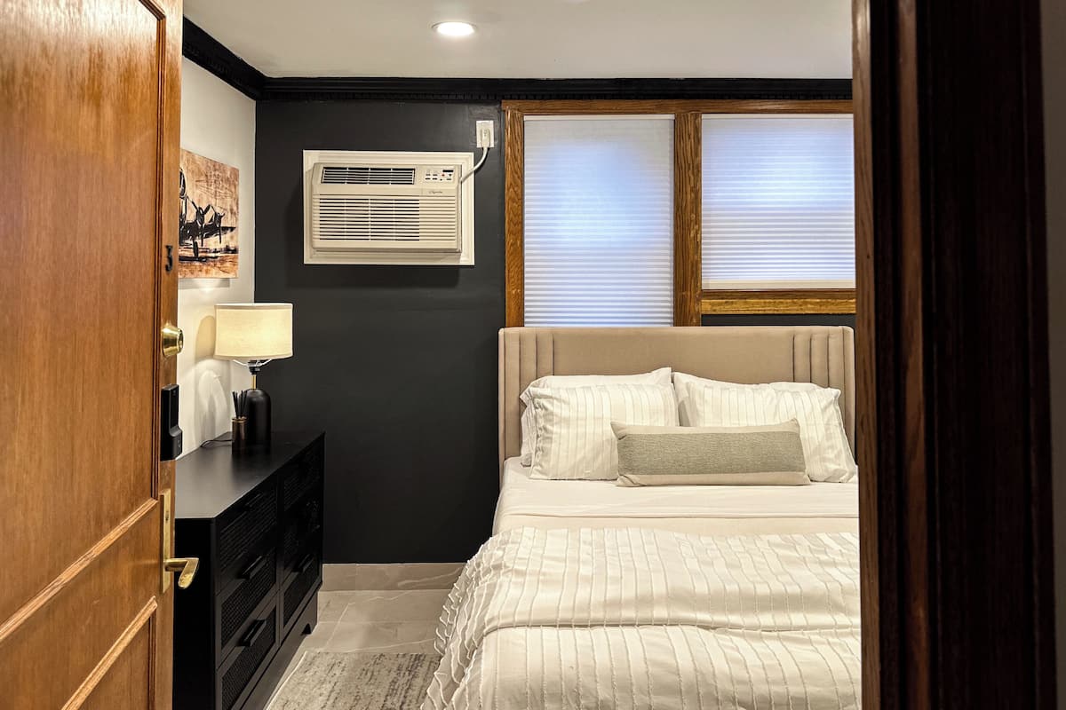 A bedroom with a bed, bedside table, lamp, air conditioning unit, and a window with blinds.