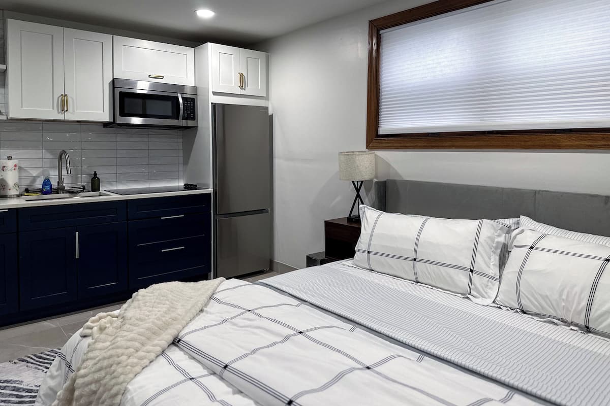 A modern hotel room with a kitchenette, a bed with striped bedding, a bedside lamp, and a window with blinds.