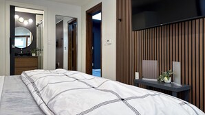 A modern bedroom with a bed, a TV mounted on a wooden panel wall, and a bathroom visible through an open door.