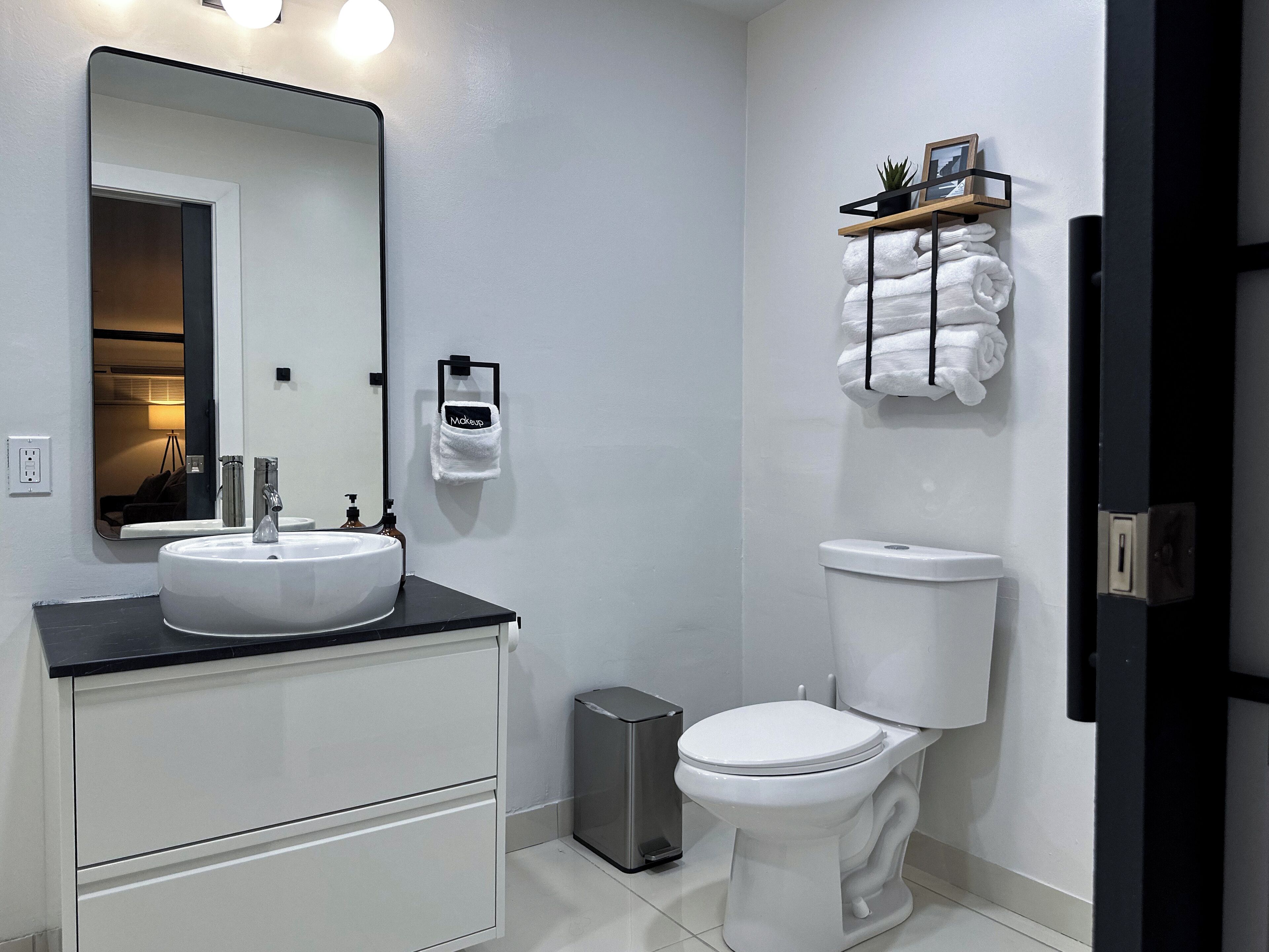 A modern bathroom with a white sink, a toilet, and a mirror. There is a trash bin and a towel rack with towels.