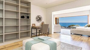 A modern living room with a large white sofa, a grey armchair, a white ottoman, a wooden desk, and a large white shelving unit.