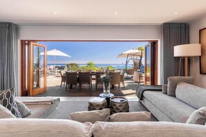 Una habitación de hotel moderna con un amplio ventanal corredero que da a un área de comedor al aire libre con sombrillas y vistas al mar.