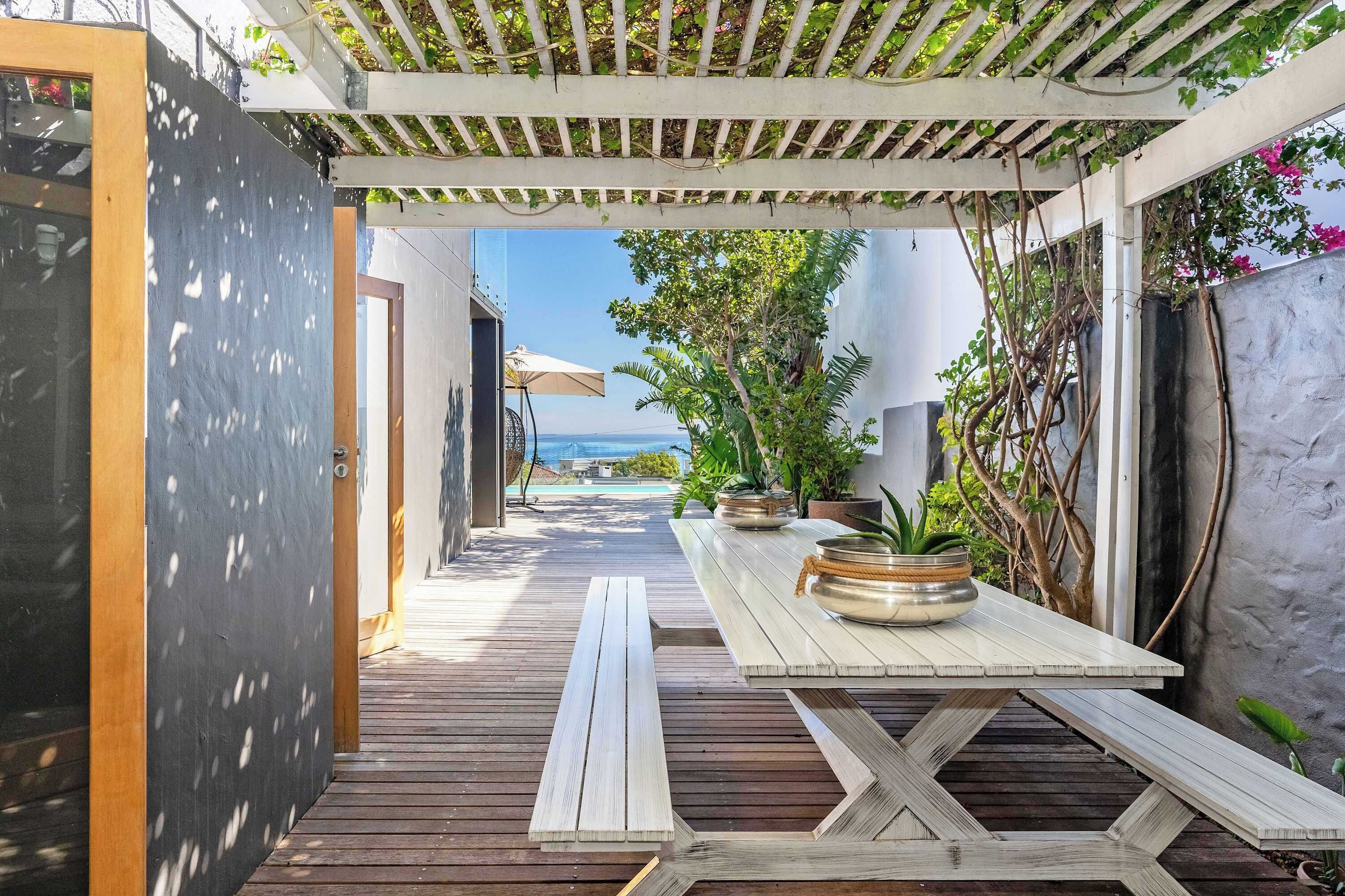 En træterrasse med et bord og stole, en pergola med slyngplanter, og havudsigt.