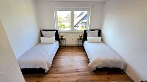 Pieni, hyvin valaistu huone, jossa on kaksi erillistä sänkyä, ikkuna, josta on näkymä rakennuksiin, ja ikkunan alla patteri.