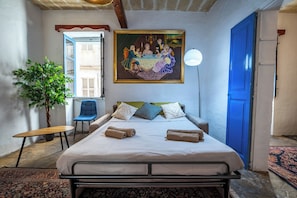 A bedroom with a bed, a chair, a table, a plant, and a painting on the wall.