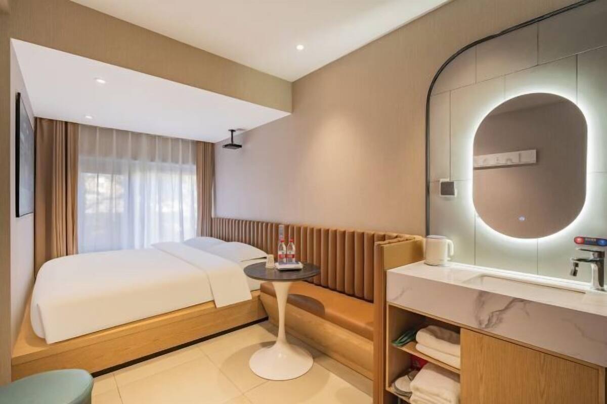Une chambre d’hôtel avec un lit, un miroir rond, une petite table avec une bouteille et une tasse, et un coin salon.