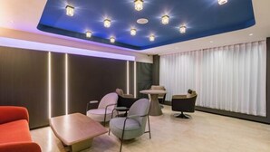 Ein modernes Hotelzimmer mit blauer Decke, integriertem Licht, einer Sitzecke mit Tisch und Stühlen sowie einem großen Fenster mit Vorhängen.