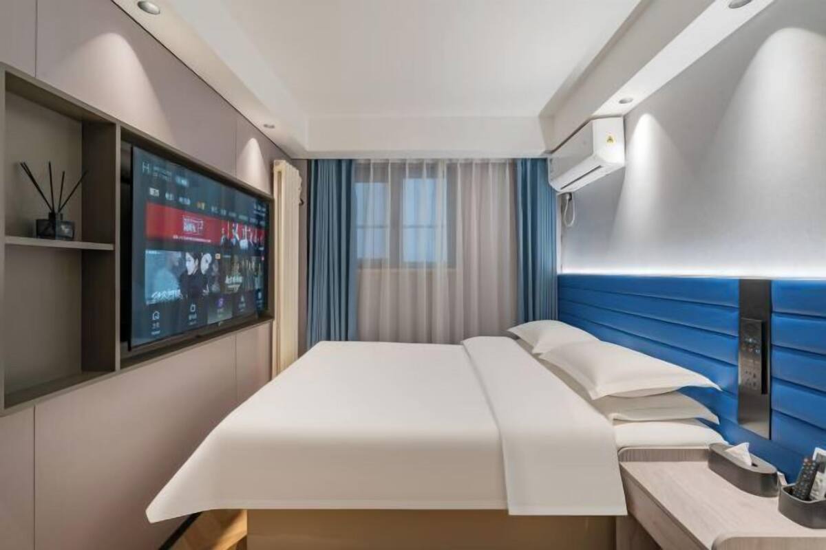 Une chambre d’hôtel moderne dotée d’une grande télévision à écran plat, d’un lit avec du linge de lit blanc, d’une table de chevet avec une télécommande et d’une tête de lit bleue.